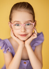 Portrait of a little girl wearing a lilac frilly dress on a yellow background. She sits, leaning on her elbows, and gently touches her face with her fingers. The model glasses with pink frames