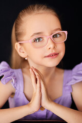 Close-up portrait of a charming young girl in pink glasses and a purple frilly dress posing on a black background. She has her palms folded together. Her hair is tied in a tight ponytail at the back.