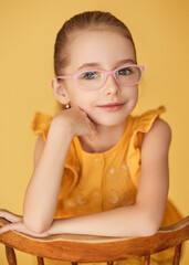 Close up of young girl with blonde hair pulled back wearing yellow ruffled top. She sits leaning on her arm and smiles tenderly. The model with expressive eyes wears corrective glasses with pink frame