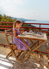 A little girl in a lilac dress is busy painting stones. She's focused on her work. Behind her, there is a beautiful view of the sea and hills. The atmosphere looks calm and peaceful