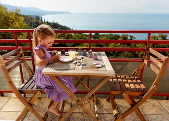A young girl with braided hair draws on rocks at a wooden table on an outdoor terrace overlooking a picturesque seascape, wooded hills. A child with glasses is engaged in creative work during summer