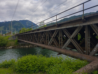 Obraz premium A sturdy metal bridge spans a flowing river, with scenic mountains in the distance, under a cloudy sky