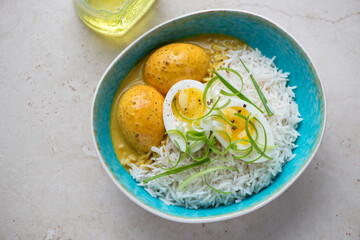 Curried eggs with rice in a turquoise bowl, horizontal shot on a light-beige stone background, high angle view