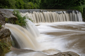 A muddy waterfall roars after recent heavy rains, captured in long exposure. The silky brown water...