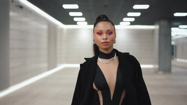 Tracking gimbal shot confident mixed race woman fashion model walking subway hall. Portrait of female wearing black bodysuit and trench coat. Futuristic architecture, sexy woman looking at camera