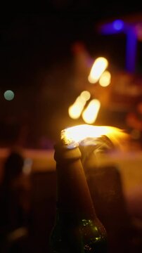 A close-up view of a flaming bottle during a dynamic fire performance at night, featuring blurred lights and a performer juggling fire in the background.