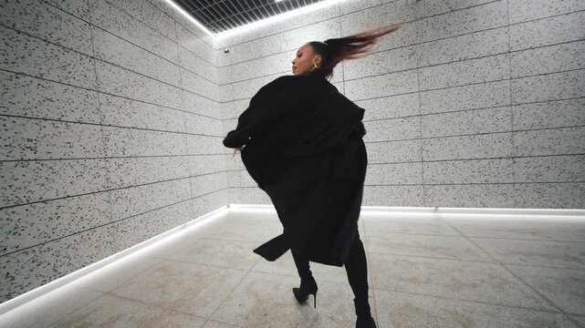 Confident black skin female dancer in sexy bodysuit, thigh high boots and long coat performs seductive choreography in futuristic underground hall with minimalistic concrete and LED lighting design