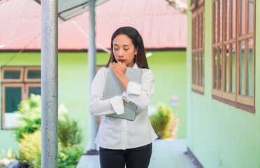 Fototapeta premium A young woman stands still, holding a laptop with a thoughtful expression. She appears to be thinking or planning, making this image ideal for concepts like decision-making, focus, or problem-solving.