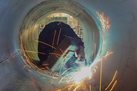 Welding inside a metal pipe. Pipeline construction in oil and gas projects. An experienced welder works inside a large metal pipe, creating bright sparks and intense light during welding. - Powered by Adobe