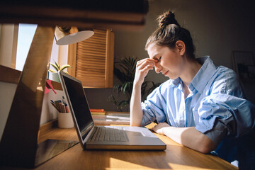 A tired young woman is sitting in front of a laptop and suffering from a headache. A freelancer...