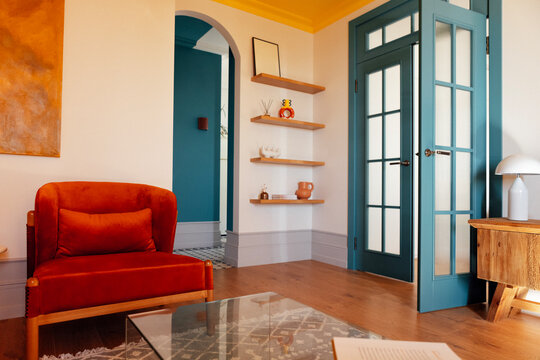A stylish living room with elegant blue doors and an arched passage. A soft orange armchair and a glass coffee table. White walls and a bright yellow ceiling.