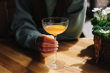 Woman holding cocktail glass. Elegant drink with orange slice. Bright natural light. Wooden table background. Stylish manicure detail. Refreshing citrus beverage. Cozy indoor setting.