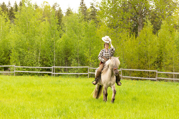 Cowgirl riding on horse (Icenlandic horse). Green meadow and trees