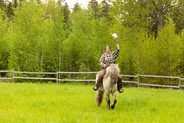 Cowgirl riding on horse (Icelandic horse). Green meadow and trees