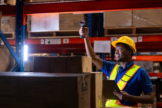 Man African American professional worker wearing safety uniform and white hard hat using bar code reader scanning box inspect product on shelves in warehouse.