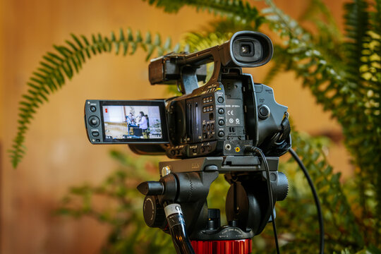 A professional video camera on a tripod records a live indoor performance with musicians and microphones, capturing the scene through its screen display.