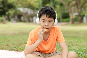 Fototapeta premium Happy Asian school boy with headphones eating snack while sitting on mat on green grass. Joyful kid spending time outdoor in garden on sunny day. Child having a picnic in summer park.