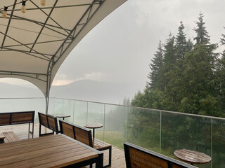 A mountaintop cafe with wooden tables and benches during a heavy rain in the mountains. Rain drops dripping down the glass fence. Silhouettes of mountains and forest in the fog in the background.