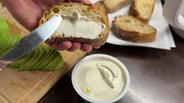 A knife spreads cream cheese on fresh crispy bread. Sliced green avocado lies below on the cutting board. A close-up of tasty and healthy breakfast preparation.