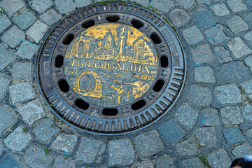 Decorative Manhole Cover Featuring Cityscape and Historical Elements on Cobblestone Street