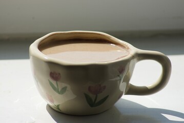 A charming ceramic cup with a light brown beverage, likely tea or coffee, sits on a sunlit windowsill. Adorned with delicate pink flowers, it offers a cozy, handmade aesthetic.