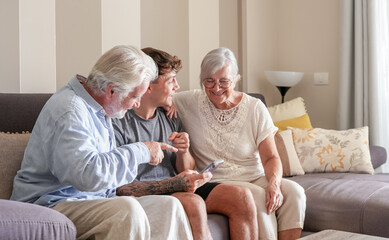 Happy Multi generation family of grandparents and young grandson looking at something funny on nephew's mobile phone sitting together at home. Old and new generation share technology