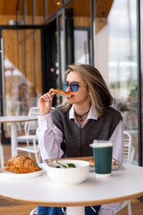 In a stylish cafe, a young woman with sunglasses enjoys a delicious meal, savoring chicken while seated at a bright white table. Sunshine pours in through large glass windows