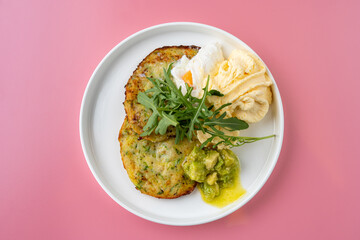 Savory potato pancakes sit elegantly on a round plate, adorned with fresh arugula and a dollop of creamy avocado. The cheerful pink background enhances the vibrant brunch experience