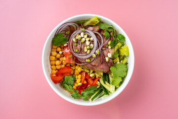 A vibrant bowl filled with fresh greens, grilled meat, sweet corn, and diced tomatoes sits atop a pastel pink surface. It showcases a mix of textures and colors, inviting anyone to dig in