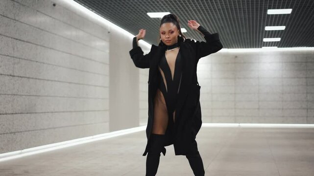 Confident mixed race woman fashion model dancing seductively and looking at camera in futuristic design underground subway hall. Female in black bodysuit, thigh-high boots and coat, concrete wall