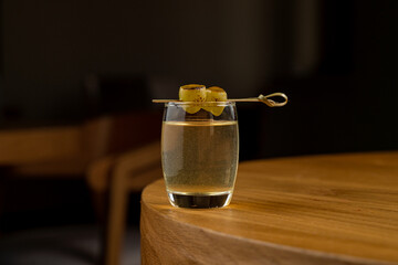 A refreshing craft cocktail rests on a minimalist wooden table, illuminated by warm ambient lighting. Its delicate garnishes of olives add a touch of elegance to the setting