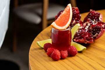 A vibrant drink featuring raspberries and citrus sits elegantly on a wooden table, surrounded by fresh pomegranate and lime slices. The warm ambiance invites relaxation and enjoyment