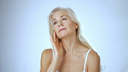 A mature woman with long silver hair smiles joyfully while gently touching her cheek in a bright...