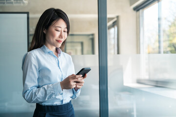 Asian young business woman manager using cell phone mobile application standing in office. 40s...