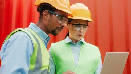 Close-up portrait of two professional workers of construction company using laptop wearing safety glasses and helmets. Young beautiful woman engineer and African male manager near red containers.
