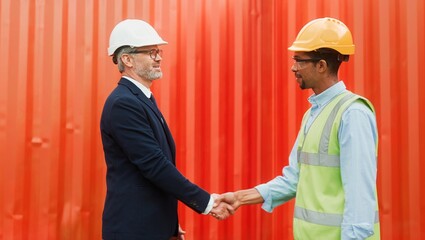 Side-view of African-American worker of transportation company talking shaking hands with ceo boss in suit. Two profesional people wearing helmets standing on ship in front of red container.