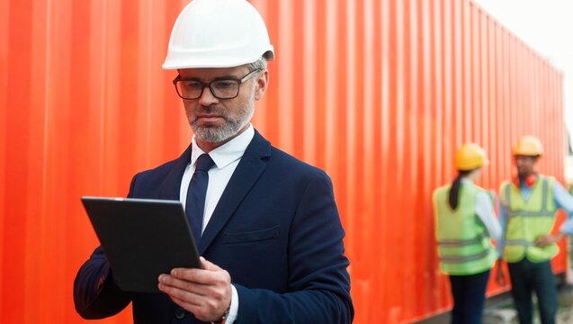 Portrait of professional thoughtful owner of shipping company using tablet working online. Handsome elegant middle-aged successful ceo in helmet and suit standing in front of container and workers.