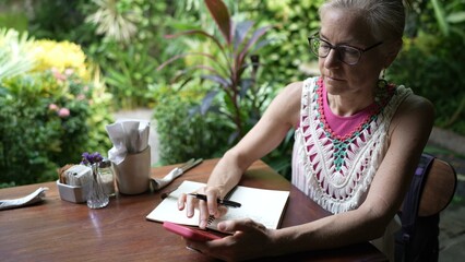 A mature woman sits at a cafe table, focused on her phone while taking notes. Surrounded by lush greenery, she enjoys her work as a digital nomad on vacation.