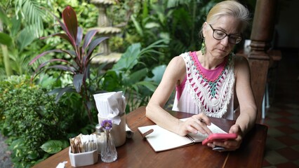 A mature woman sits at a cafe table, writing in a notebook and using her phone, surrounded by lush greenery and a relaxed atmosphere.