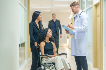 sick african business woman sitting on wheelchair at corridor with asian woman assistant and colleagues walking to encourage her,getting service at examine room in hospital