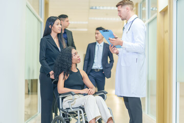 sick african business woman sitting on wheelchair at corridor with asian woman assistant and colleagues walking to encourage her,getting service at examine room in hospital