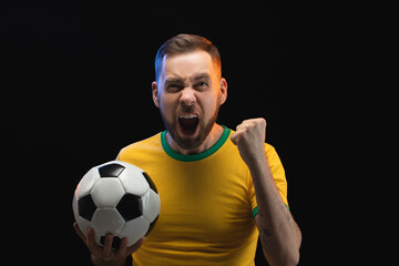 Studio shot of euphoric soccer player or supporter man in yellow t-shirt is holding ball in hand, screaming and making winner's gesture "yes" clenching his fist