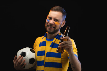 Studio shot of joyful soccer supporter wearing yellow-blue uniform is celebrating his favourite team win, raising beer bottle while posing with a ball in hands, isolated over black background