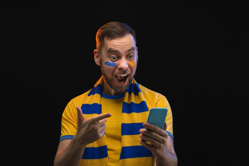 Studio shot of euphoric soccer supporter pointing at his phone screen with index finger celebrating triumph, his bet played, isolated over black background