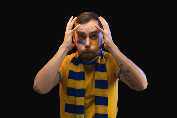Studio shot of emotional soccer supporter man in yellow-blue t-shirt and scarf with painted face, looking nervous, holding his head in despair while watching play, isolated on black background