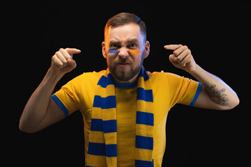 Studio shot of emotional soccer supporter man with painted face, wearing yellow-blue t-shirt and scarf, looking furious, ponting with index fingers at the camera, isolated on black background