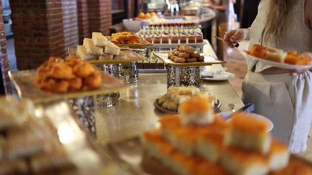 Middle Eastern dessert buffet woman picking kunafa baklava sweet pastries vibrant colors golden textures indulgent variety cozy atmosphere rich flavors inviting hotel dining experience