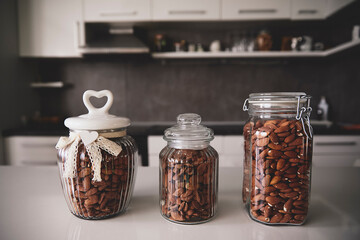 A trio of glass jars filled with almonds on a kitchen counter. Elegant presentation with heart shaped lid decor and modern kitchen backdrop. Healthy food and lifestyle concept