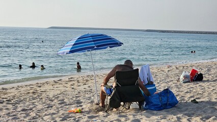 A serene beach scene featuring a person relaxing under a large umbrella, seated in a comfortable paper chair while soaking up the sun. Nearby, several packages rest on the sand, adding to the tranquil