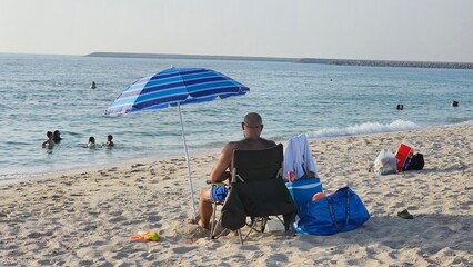 A serene beach scene featuring a person relaxing under a large umbrella, seated in a comfortable paper chair while soaking up the sun. Nearby, several packages rest on the sand, adding to the tranquil